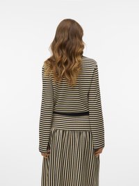 Donna con lunghi capelli ondulati, che indossa un top a maniche lunghe beige e nero a righe e una gonna plissettata coordinata, di spalle su uno sfondo bianco.