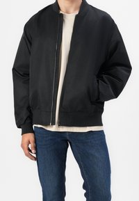 Chaqueta bomber negra con una textura suave, cierre de cremallera, cuello y puños acanalados. Llevada sobre una camiseta de color claro y unos vaqueros azules.