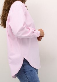 Chemise rose pâle à manches longues avec un ourlet arrondi, tissu en coton doux, poignets boutonnés et coupe décontractée, portée avec un jean bleu.