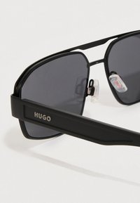 Čierne slnečné okuliare s tmavými obdĺžnikovými sklami a brandingom "HUGO" na stranke, vybavené nastaviteľnými priehľadnými nosovými podložkami.
