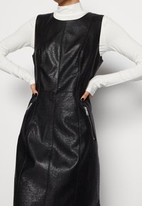 Schwarzes Lederkleid ohne Ärmel mit glatter Textur, ausgestattet mit zwei seitlichen Reißverschlusstaschen und ausgeprägten Nähten entlang des Oberteils.