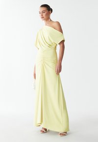 Robe jaune à une épaule avec un tissu drapé, des détails froncés à la taille et un ourlet longue. Le mannequin pose avec un sac à main transparent et des talons.