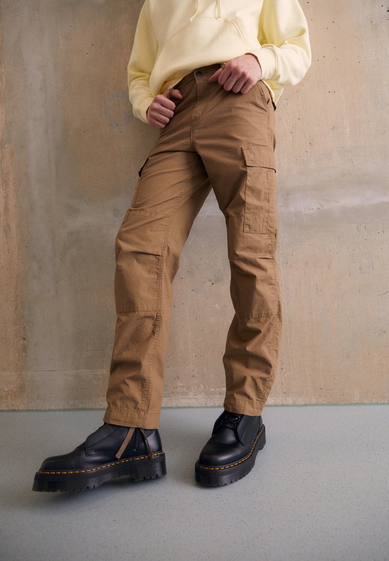 Personne portant un pantalon cargo beige, des bottines noires épaisses à lacets et un hoodie jaune pâle, debout devant un mur beige uni.