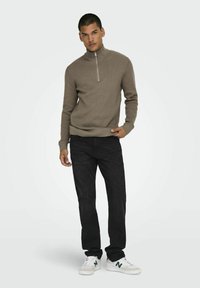 Brown knit sweater with a high collar and zip closure, paired with black jeans and white sneakers with green accents.