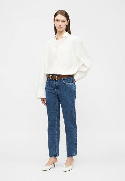 Woman standing with hand in pocket wearing white oversized shirt, blue jeans with brown belt, and white heels on a plain background.