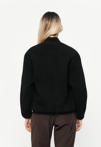 Must must jacket, mille tekstureeritud pinnaga, ümmarguse krae ja laia lõikega. Ühendatud pruuni pükstega, tagant vaates ei ole lisadetailide.