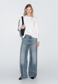 Weißes Langarmshirt, hellblaue weite Jeans, hält eine schwarze Tasche. Weicher Stoff, lässiges Design mit lockerer Passform und geradem Bein.