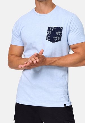 Mężczyzna w jasnoniebieskim t-shircie z krótkim rękawem i granatową kieszenią na piersi z białymi wzorami ptaków, splatający ręce na wysokości talii.
