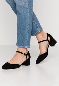 Zapatos de tacón con tira de tobillo de ante negro, con punta afilada y tacón cuadrado. Las tiras tienen un detalle de hebilla dorada. Llevados con jeans azules acampanados.