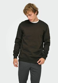 Jeune homme portant un sweat-shirt noir à manches longues et un pantalon gris foncé, regardant vers le bas tandis que sa main droite ajuste l'ourlet du sweat-shirt.