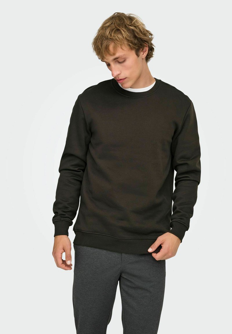 Jeune homme portant un sweat-shirt noir à manches longues et un pantalon gris foncé, regardant vers le bas tandis que sa main droite ajuste l'ourlet du sweat-shirt.