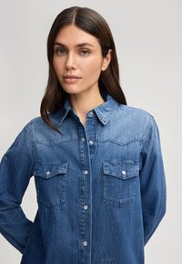 Camicia di denim di un blu medio con colletto a punta, due tasche sul petto, bottoni a scatto e design a yoke in stile western.