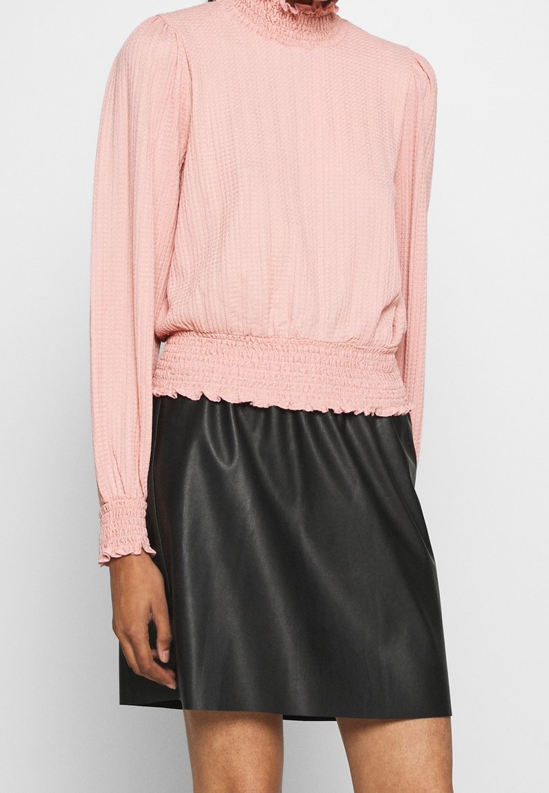 Top lungo manica textured color rosa con collo alto arricciato, abbinato a una gonna nera in finta pelle liscia. Dettaglio di vita arricciata.