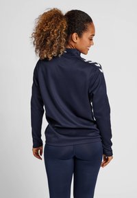Veste de sport marine avec col haut, manches longues et accents blancs en chevron sur les épaules. Tissu lisse et extensible. Leggings marins assortis.