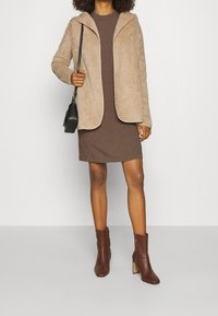 Veste en fausse fourrure beige avec capuche, robe marron à côtes et bottines marron à talon bloc. Un sac bandoulière noir complète le look.