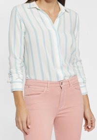 White button-up shirt with blue vertical stripes, featuring long sleeves and a collar, paired with pink fitted jeans.