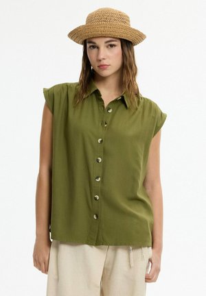 Mujer con sombrero de paja, camisa sin mangas verde oliva abotonada y pantalones beige, de pie frente a un fondo claro y liso.