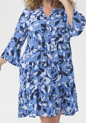 Blue, white, and black abstract floral knee-length dress with a V-neck and three-quarter bell sleeves worn by a curly-haired person.