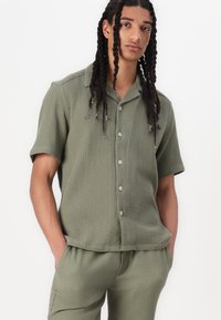 Camisa de manga corta, de color verde oliva, acanalada, con un cierre de botones en la parte delantera, cuello recto y corte relajado. Se lleva con pantalones texturizados a juego.