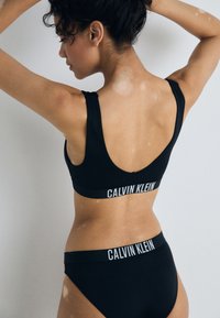 Crna sportska brusnica i odgovarajući tangice s bijelim logotipom "CALVIN KLEIN". Ima široke naramenice i elastičnu traku u struku, s glatkom teksturom tkanine.