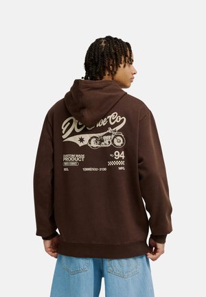 Person in braunem Hoodie mit Vintage-Motorrad-Grafik und Text auf dem Rücken, trägt hellblaue Jeans und ist mit dem Rücken zur Kamera.