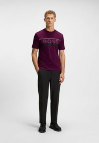 Lila idrottströja med "BOSS"-logotyp, svart accentrand, ihop med svarta skräddarsydda byxor och svarta sneakers, stående framför en neutral bakgrund.