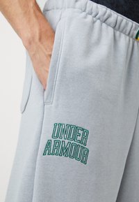 Světle šedé tepláky s elastickým pasem, bočními kapsami a zeleným vyšitým logem "Under Armour" na stehně. Měkká textura.