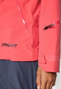 Rote wasserdichte Jacke mit einer Reißverschlusstasche, verstellbarem Handgelenkverschluss mit Klettverschluss und schwarzem Logo. Glatte Textur mit tailliertem Design.