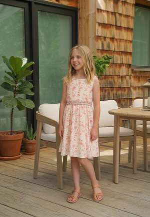 Junges Mädchen in einem geblümten Kleid und Sandalen steht auf einer hölzernen Terrasse, im Hintergrund sind Topfpflanzen und Gartenmöbel zu sehen.