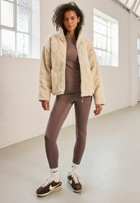 Cremiger, flauschiger Reißverschluss-Jacke mit Seitentaschen über einem braunen, taillierten Oberteil und passenden Leggings, kombiniert mit braunen Sneakern und weißen Knöchelsocken.