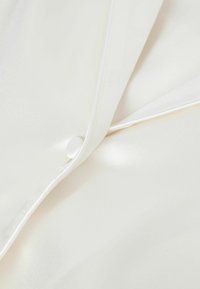 Närbild av en krämfärgad satin skjorta med rund knapp, slät tygkvalitet och jämn yta med subtil lyster; minimal sömnadsdetalj synlig.