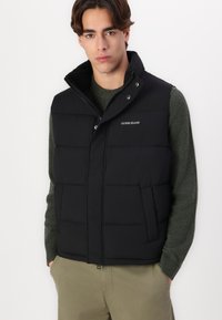 Black puffer vest with a high collar and zip closure, featuring a quilted design and logo detail on the chest. Worn over a green sweater.
