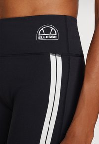 Črne legice z visokim pasom, ki imajo dve navpični beli črti in reliefni logotip na vrhu. Gladek, raztegljiv material.