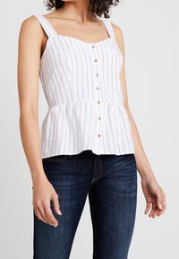 Biała bluzka peplum w niebieskie pionowe paski, kwadratowy dekolt i guziki z przodu. Gładka tekstura, szerokie ramiączka i dopasowana talia.