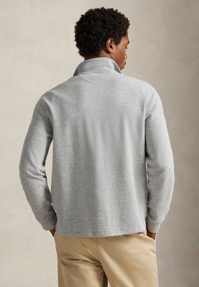 Homme portant un pull gris clair à manches longues avec col montant et pantalon beige, debout les mains dans les poches, de dos.