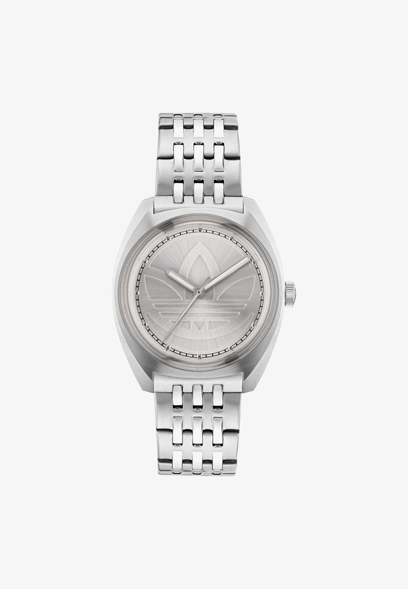 Sudraba metāla pulkstenis ar apaļu korpusu, strukturētu ciparnei un iegravētu logotipu. Iezīmēta ar saistītu siksnu un vienkāršām sudraba rādītājām.