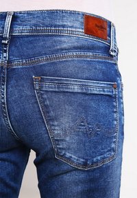 Blå denimjeans har en bakfickdesign med kontrasterande sömmar och en brun läderetikett nära midjebandet. Texturerat tyg med blekningsdetaljer.