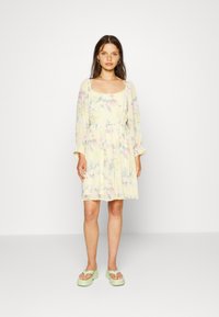 Vestido de manga curta com padrão floral em amarelo pálido, com decote quadrado e bainha com babados, combinado com sandálias de plataforma verdes.