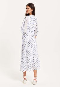 White dress with blue polka dots and floral patterns, flowing fabric, three-quarter sleeves, tied at the waist, worn with white block-heeled sandals.