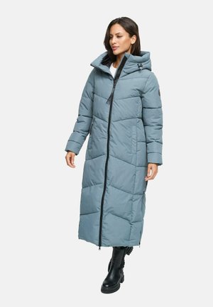 Woman wearing long blue padded winter coat with hood and black boots, standing with arms slightly raised against white background.