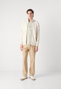 Ljus krämfärgad zip-jacka över en beige t-shirt med grafisk design, matchad med ljusbeiga byxor och vita sneakers med bruna detaljer.