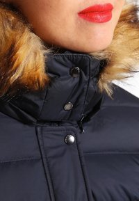 Zwarte donzen jas met een nepbontkraag in bruine tinten, voorzien van metalen drukknopen en een textuur in de stof.