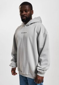 Lys grå hoodie med en frontlomme i kangaroo-stil. Har et sort logo "R&CWEAR" og ribbede manchetter og talje. Blød, afslappet pasform.