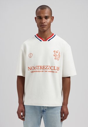 Mężczyzna noszący biały oversize T-shirt z czerwono-niebieskimi paskami na kołnierzyku, pomarańczowym napisem "NOSTREZZCLUB" i grafiką lwa, w zestawieniu z jasnoniebieskimi dżinsami.