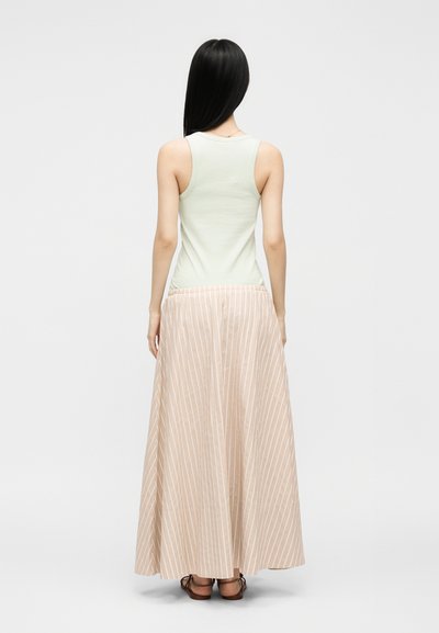 Femme aux longs cheveux noirs portant un haut sans manches vert clair et une jupe maxi beige rayée, debout de dos sur fond blanc.