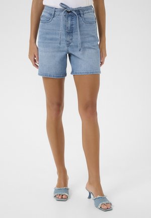 Kvinde iført lyseblå denimshorts med bundet bælte, kombineret med matchende åbne hælede sandaler.