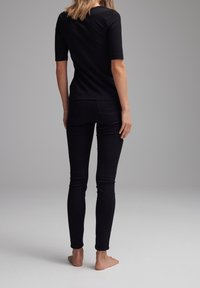 Woman standing barefoot facing away, wearing black short-sleeve top and black skinny jeans against neutral background.