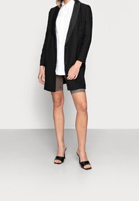 Black blazer with a satin lapel, white button-up shirt, patterned shorts, and open-toe black heeled mules. Smooth fabric and tailored fit.