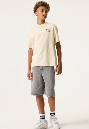 Cremefarbenes Baumwoll-T-Shirt mit dem Aufdruck "EFFORTLESS DAYS" auf der Brust, kombiniert mit grauen Jeansshorts und weißen Sneakers mit schwarzen Streifen.