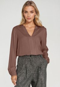 Brązowa bluzka z dekoltem V i długimi rękawami, wykonana z gładkiego materiału. Charakteryzuje się subtelnym wzorem fałd i zestawiona z szarymi spodniami w kratę.